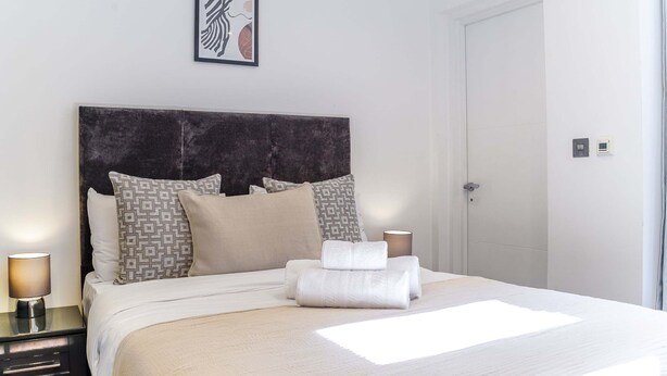 A neatly made bed with a headboard, pillows, and a rolled towel. There is a bedside lamp, a framed picture on the wall, and a door to the right. - 2BR 2BA near Hyde Park & Kensington (London)