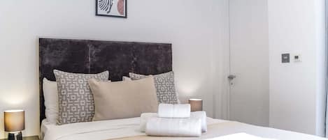 Un lit bien fait, avec une tête de lit, des oreillers et une serviette enroulée. On y voit une lampe de chevet, un tableau encadré au mur et une porte sur la droite.