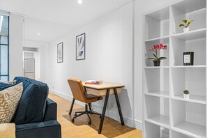 A modern living room with a dark blue sofa, a wooden desk with a chair, white shelving, and two framed artworks on the wall. - Modern 2-Bedroom in the Heart of Holborn (London)