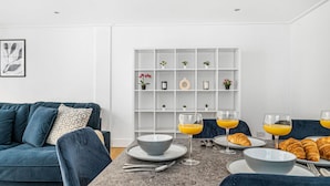 Ein modernes Wohnzimmer mit einem dunkelblauen Sofa, einem Esstisch mit Tellern, Gläsern und Croissants sowie einer weißen Regaleinheit mit kleinen Pflanzen.