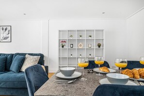 A modern living room with a dark blue sofa, a dining table with plates, glasses, and croissants, and a white shelving unit with small plants.