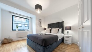 Ein modernes Schlafzimmer mit einem großen Bett, einem Nachttisch, einer Lampe und einem Fenster mit Blick auf Gebäude.