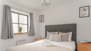 A neatly arranged bedroom with a large bed, a bedside table, a nightstand, a window with curtains, a potted plant, and a framed picture on the wall. - Spacious 2-Bedroom Flat in London's Holborn (London)
