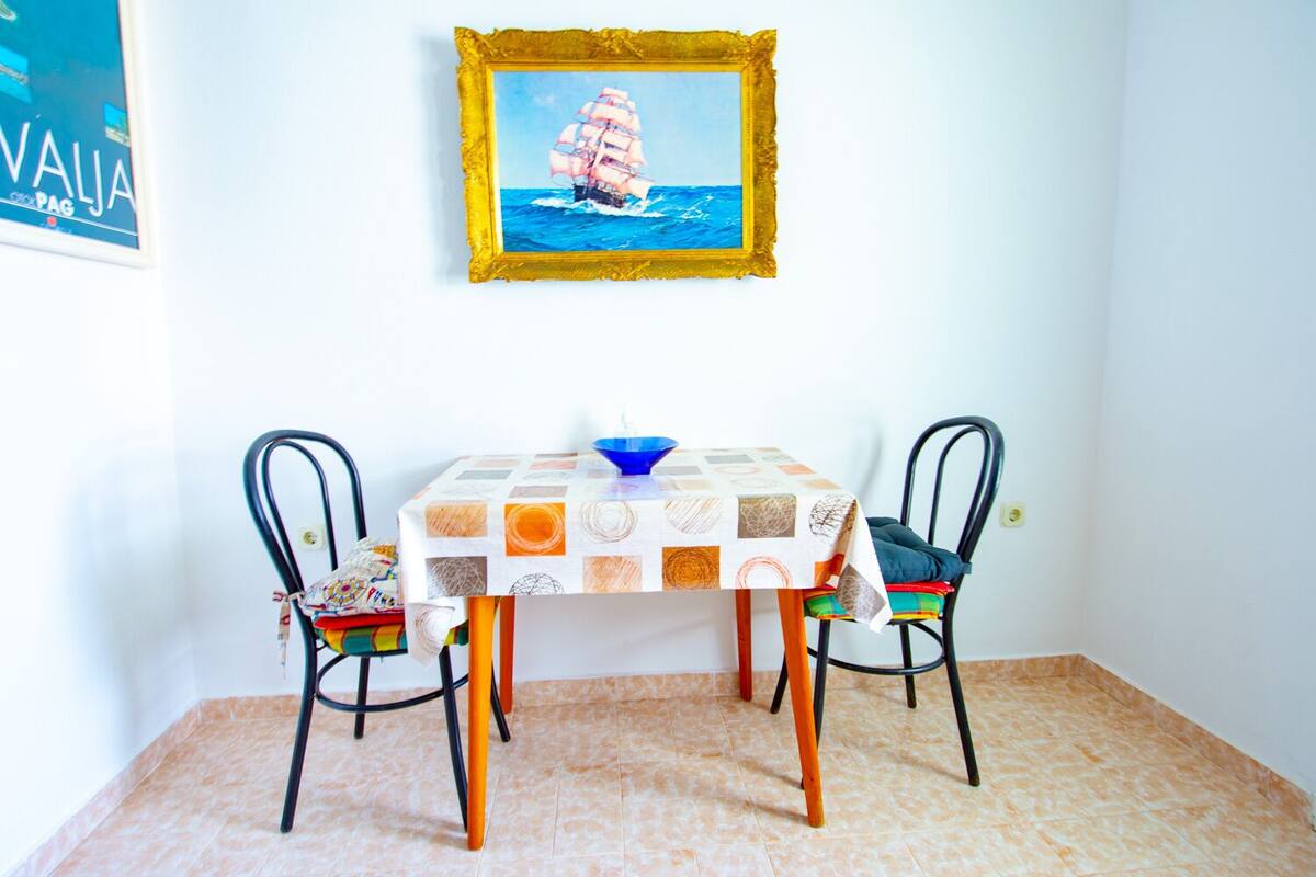 Un coin repas avec une table recouverte d’une nappe à motifs, deux chaises et un tableau encadré représentant un voilier, accroché au mur.