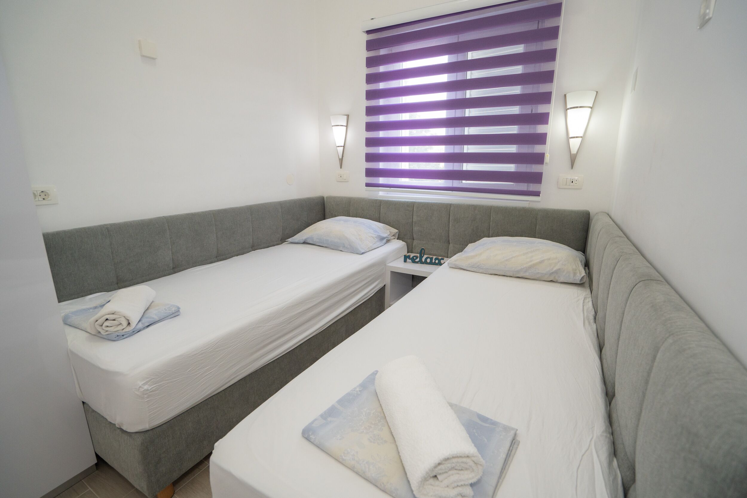 A twin room with two single beds, a small table, and a window with purple blinds.