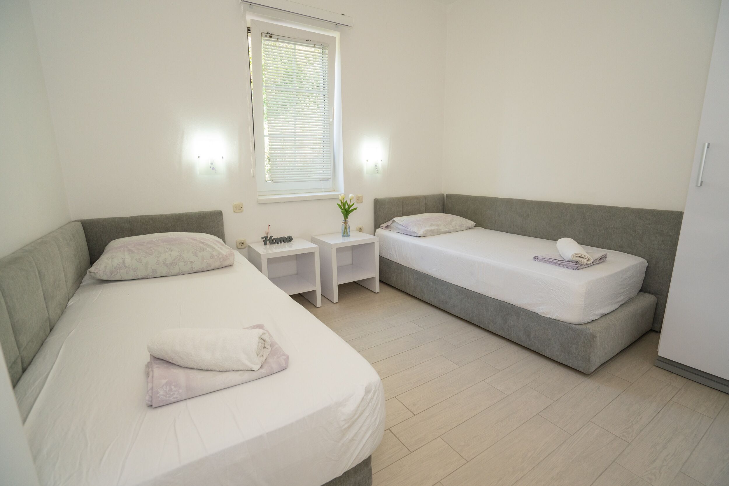 A bedroom with two single beds, a nightstand with a vase, and a window with blinds.