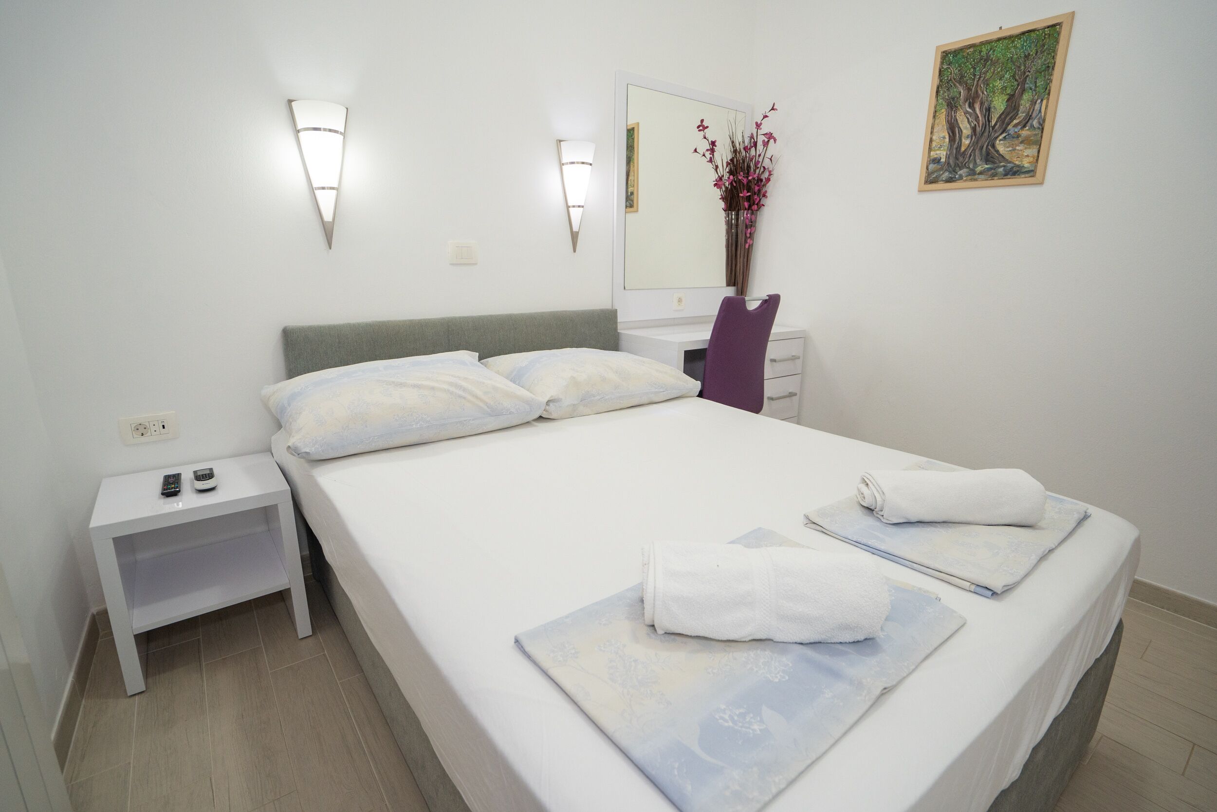 A neatly made bed with white linens, a small bedside table with a remote control, a mirror, and a framed artwork on the wall.