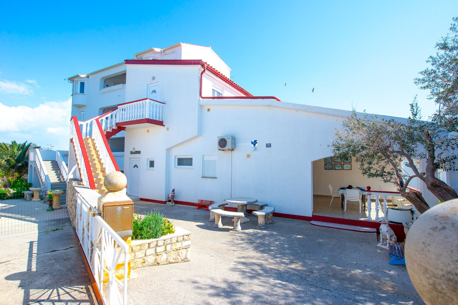 A white building with a red roof, a staircase, outdoor seating, and a tree.