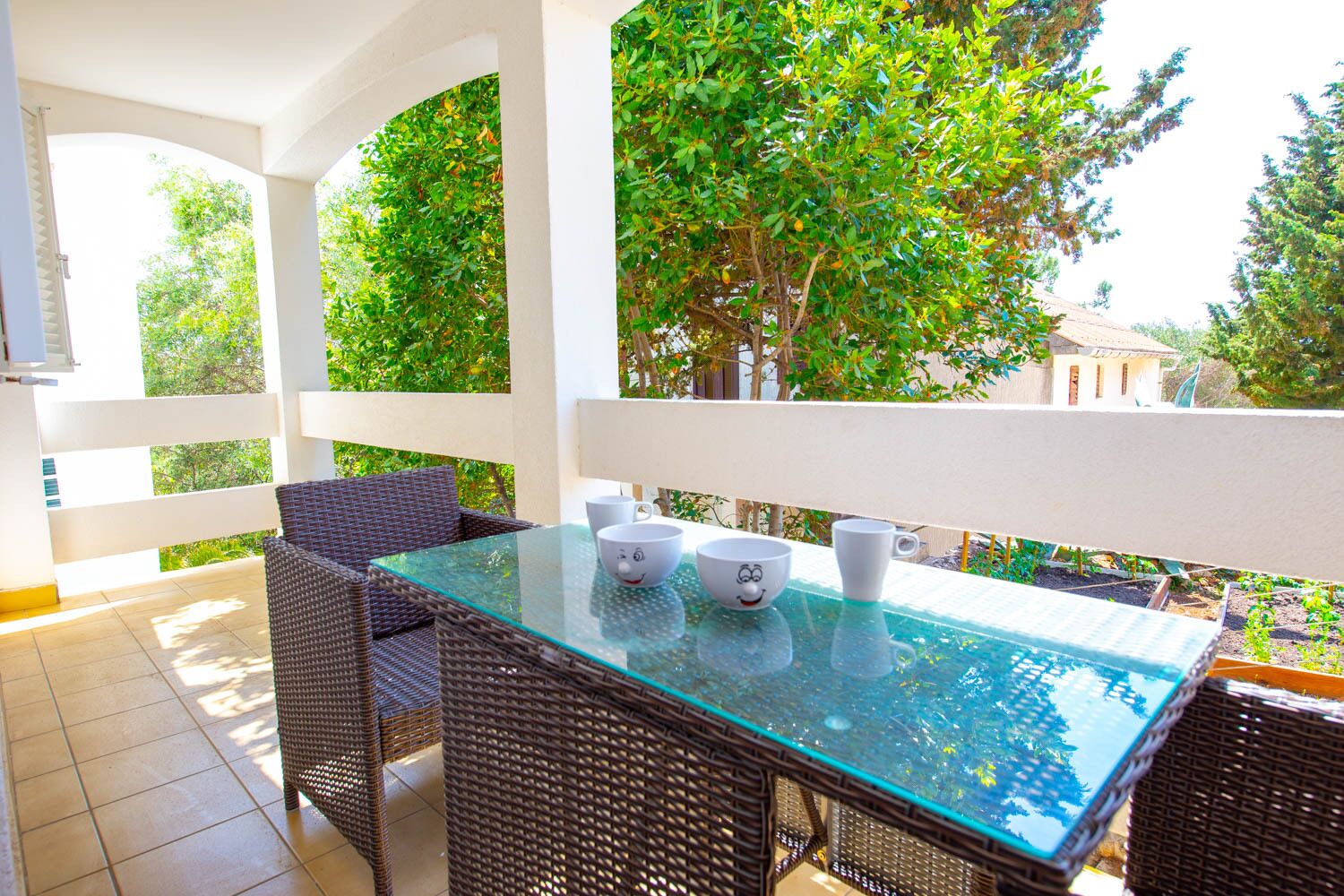 A glass-top table with wicker chairs and white mugs with faces on a balcony with a view of trees and a building.
