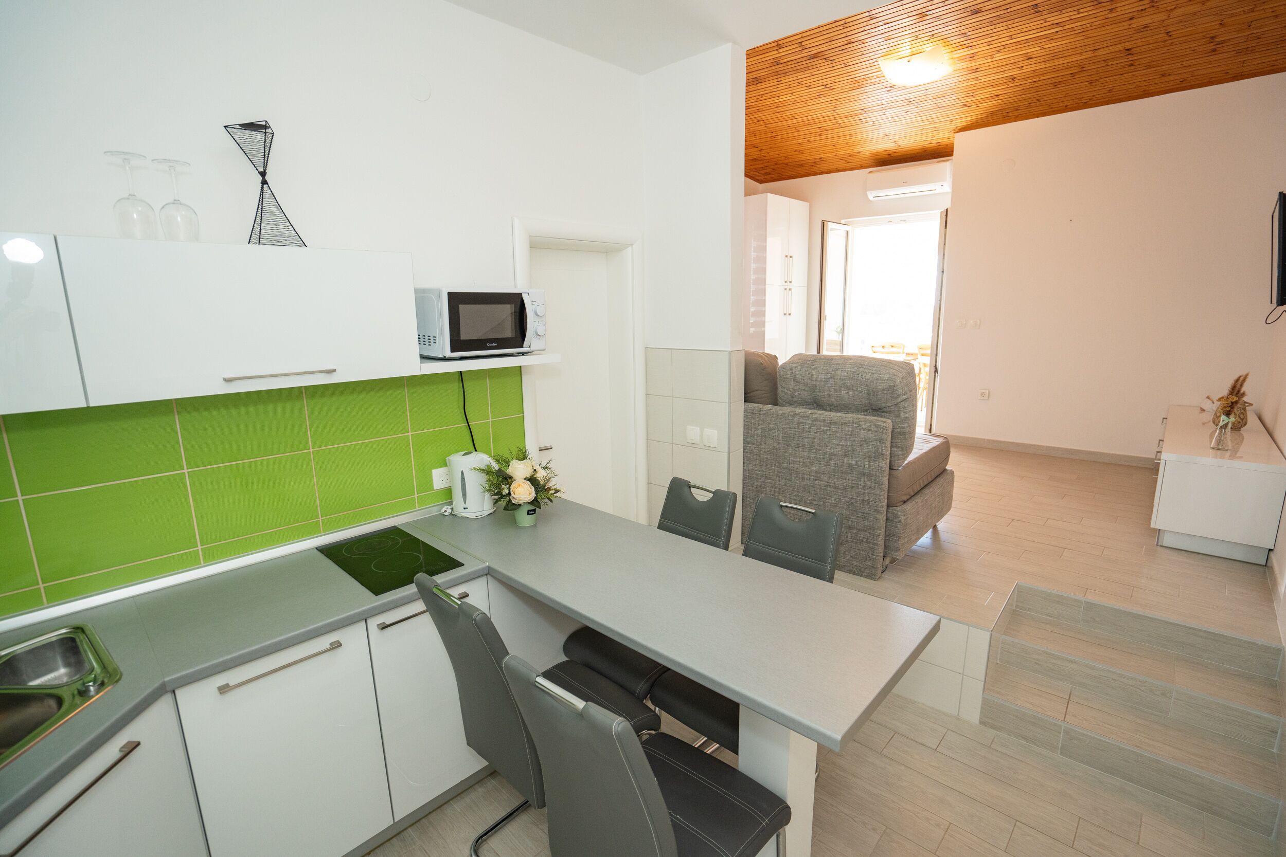 A modern kitchen with a grey countertop, white cabinets, and a green backsplash. There is a microwave on the countertop, a sink, and a chair.