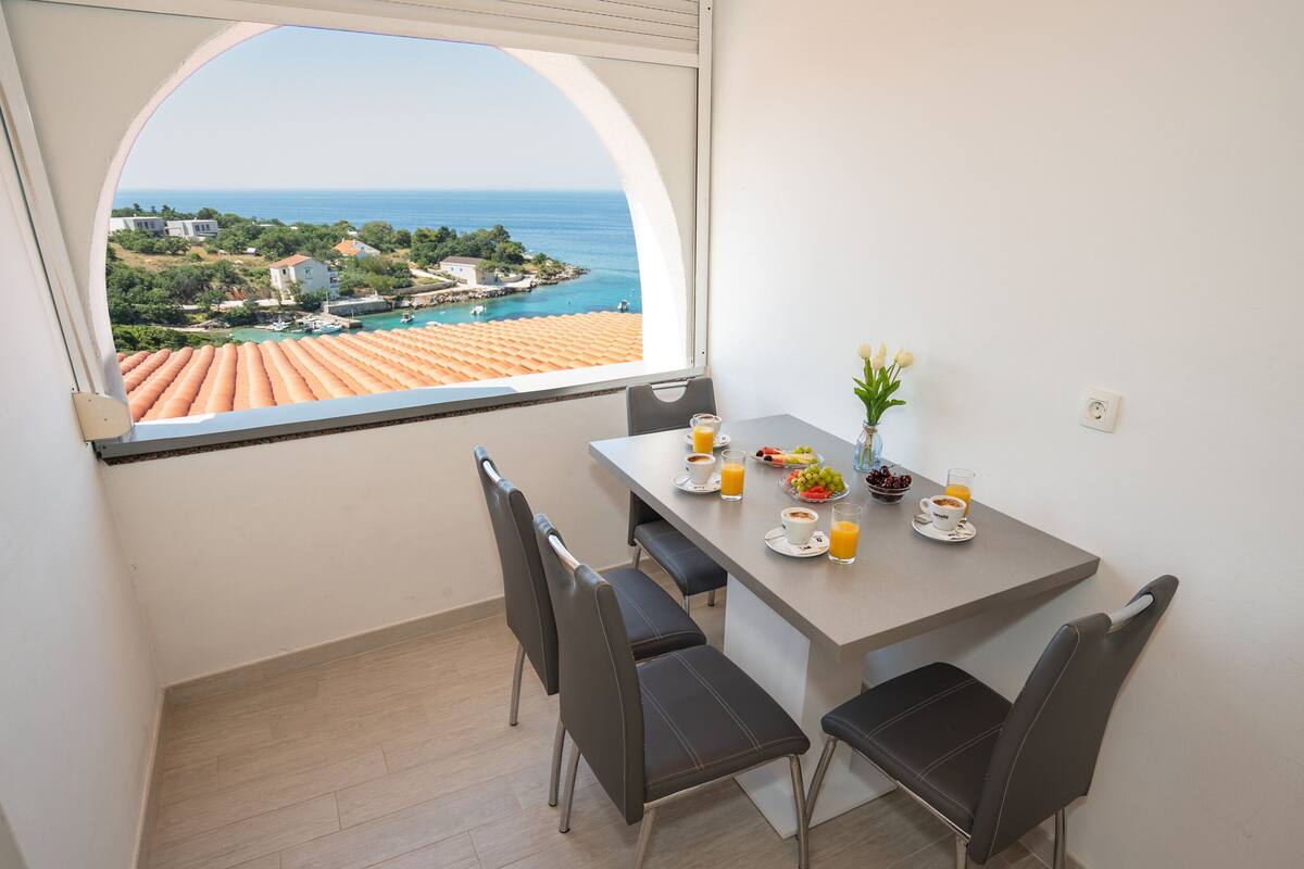 Un coin repas avec une table dressée pour deux personnes, une vue sur la mer et une fenêtre donnant sur la côte.
