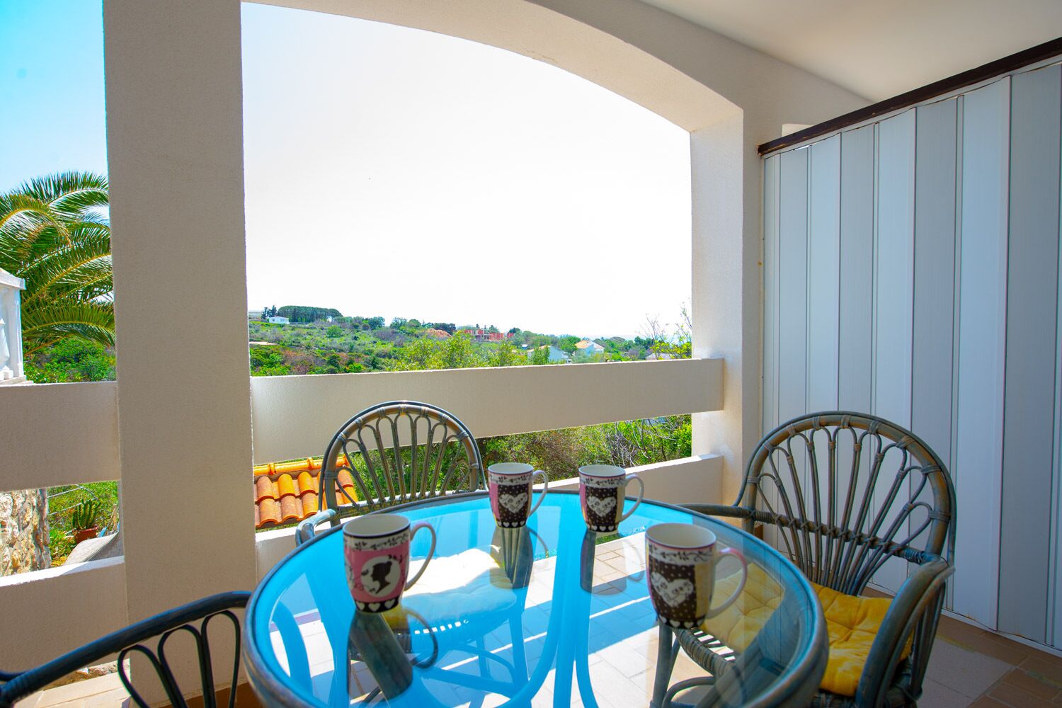 Una mesa de vidrio con cuatro sillas y cuatro tazas en un balcón que ofrece vistas a zonas verdes y edificios.