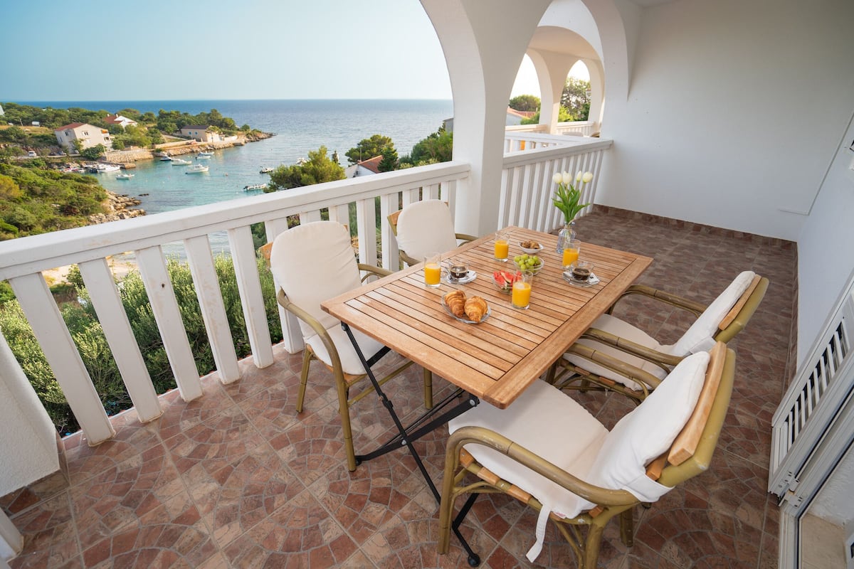 Une terrasse avec une table en bois, des chaises blanches dotées de coussins et une vue sur la mer.