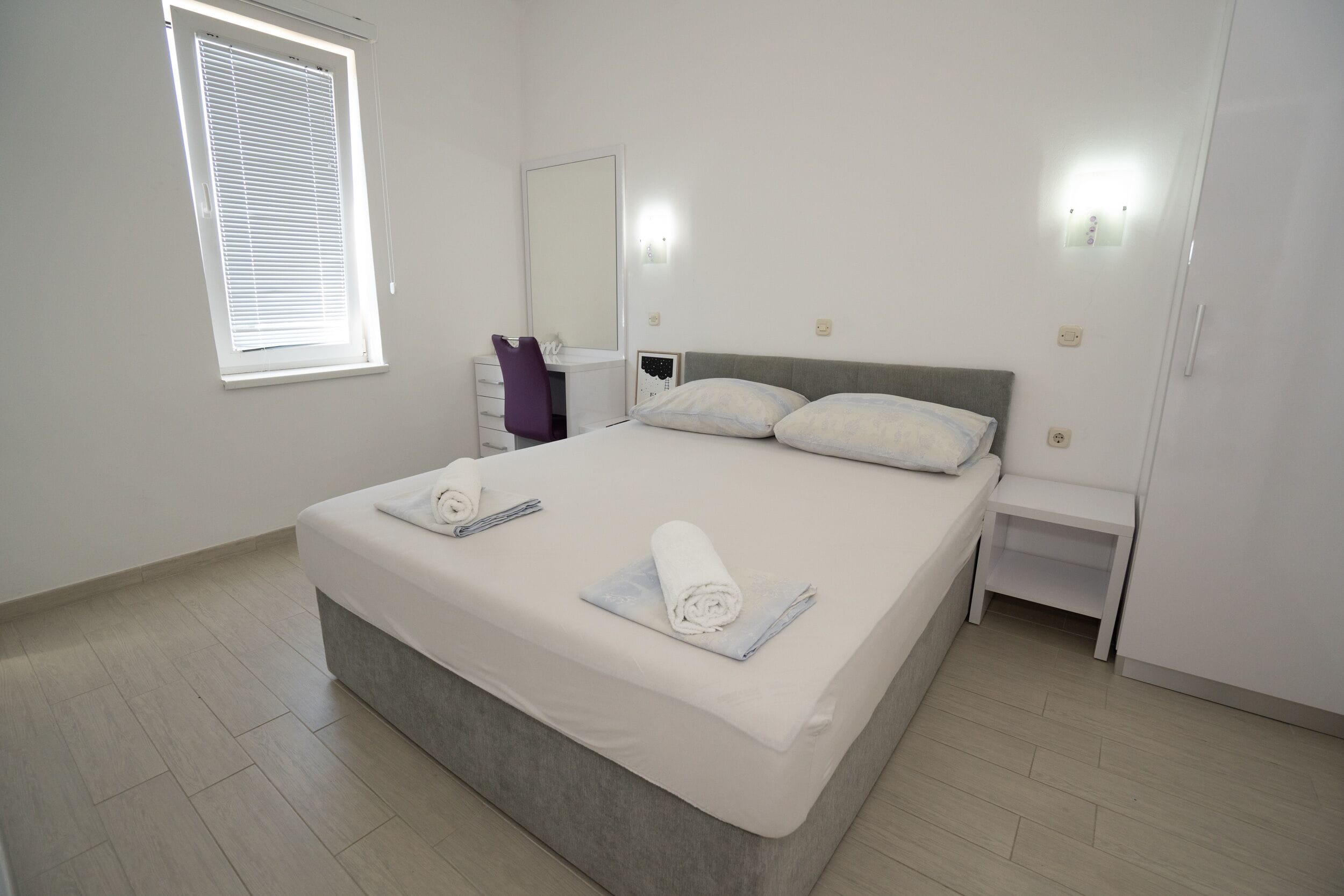 A bedroom with a large bed, a bedside table, a chair, a mirror, and a window with blinds.