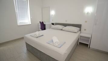 A bedroom with a large bed, a bedside table, a chair, a mirror, and a window with blinds.
