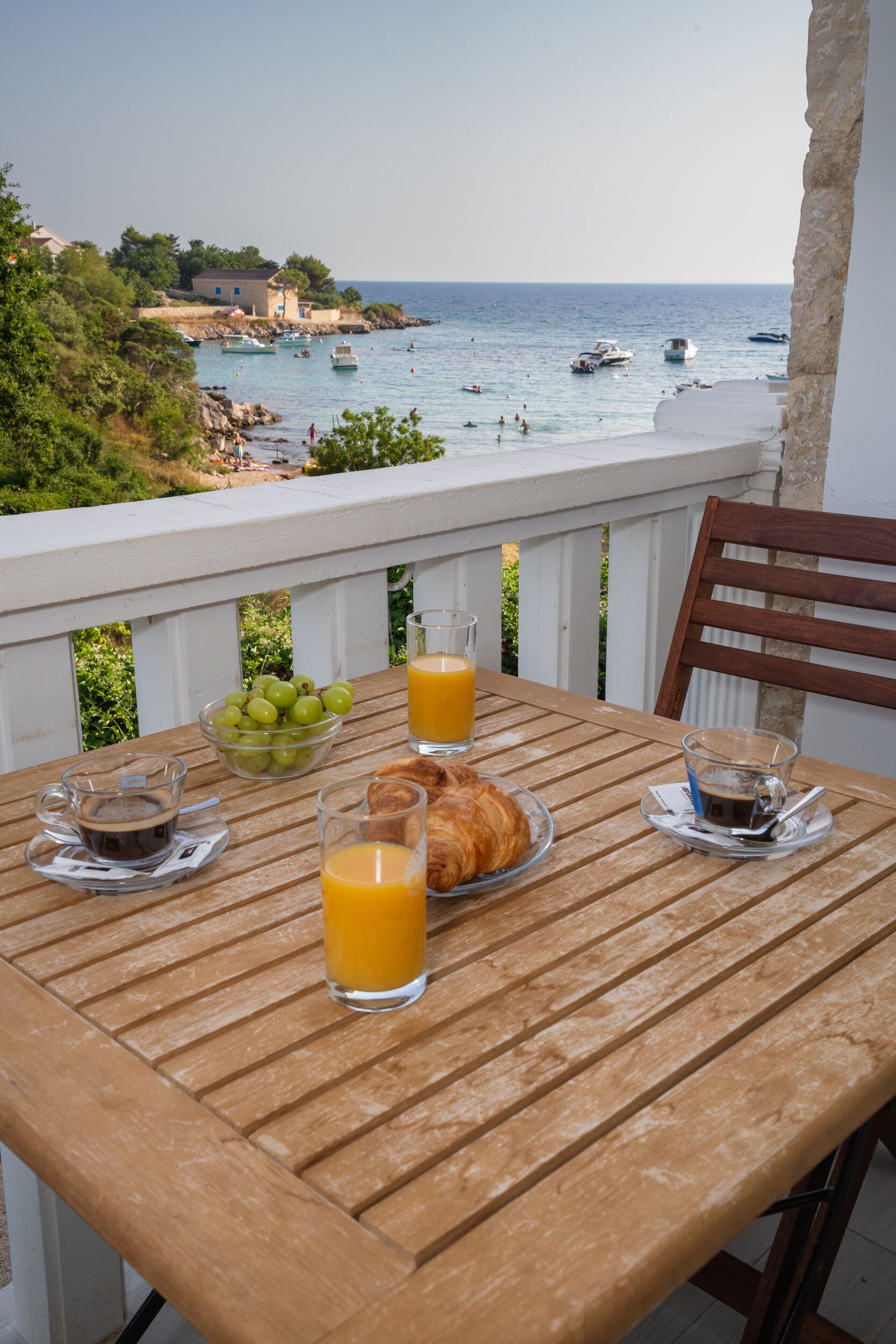 A wooden table with breakfast items and a glass of orange juice, overlooking a coastal area with boats and greenery.
