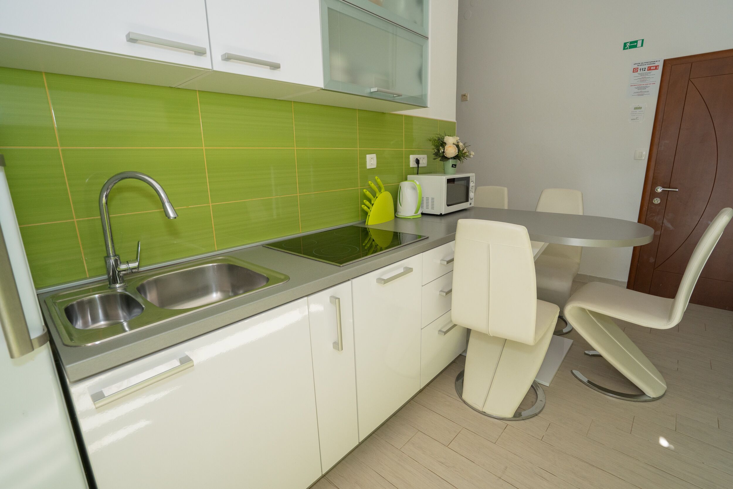 Una cocina moderna con armarios blancos, un salpicadero verde y un lavamanos de acero inoxidable. Hay un microondas, una mesa pequeña con sillas y una puerta con un letrero de salida de emergencia.