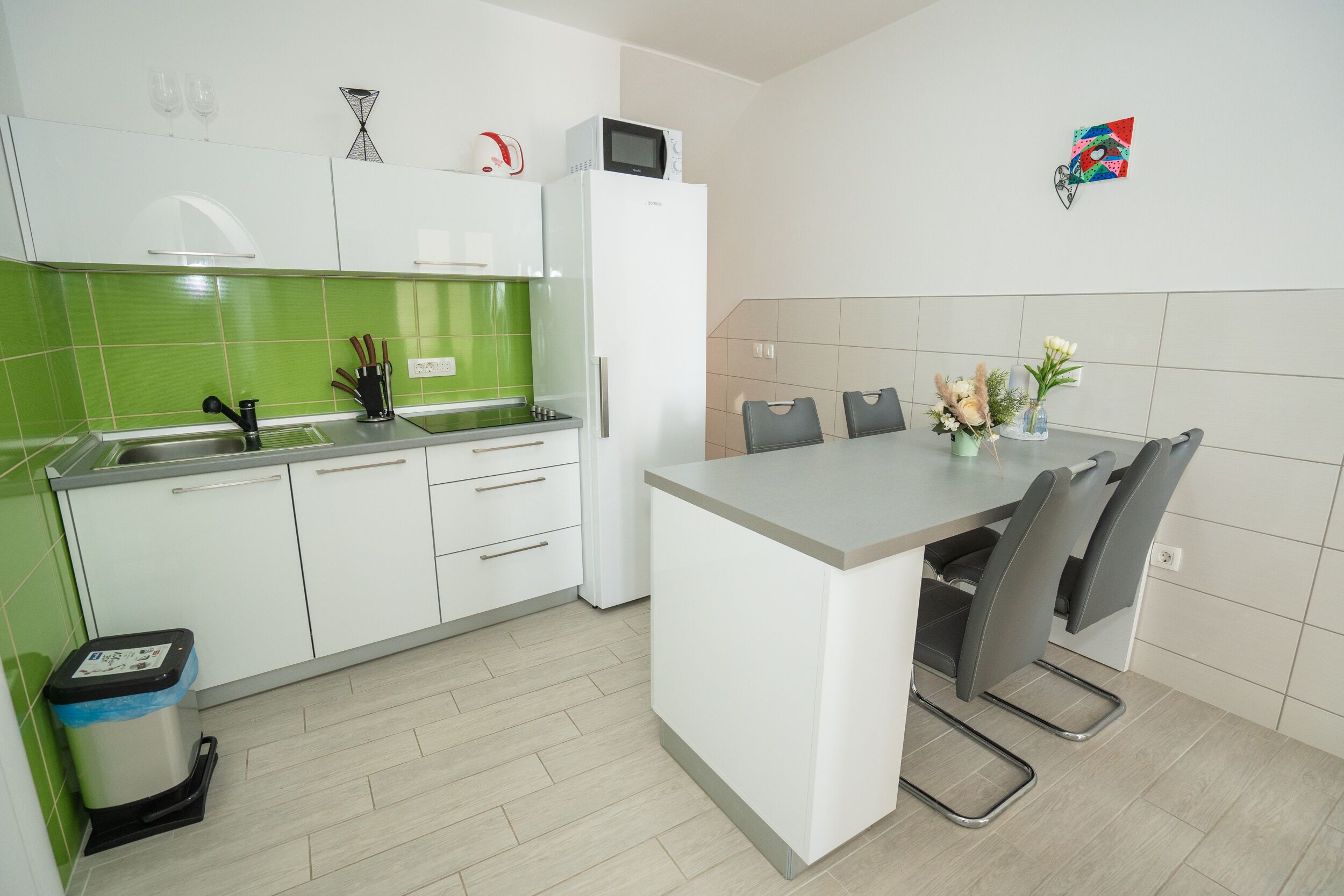 A modern kitchen with white cabinets, a green backsplash, a microwave, and a dining area with chairs.