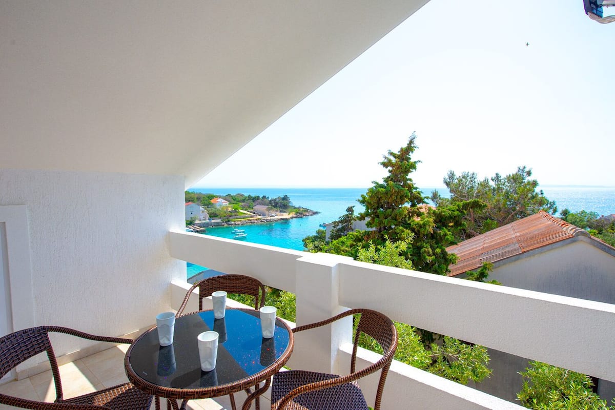 Un balcon avec des fauteuils en osier et une table, offrant une vue sur la côte, avec des bâtiments et des arbres.