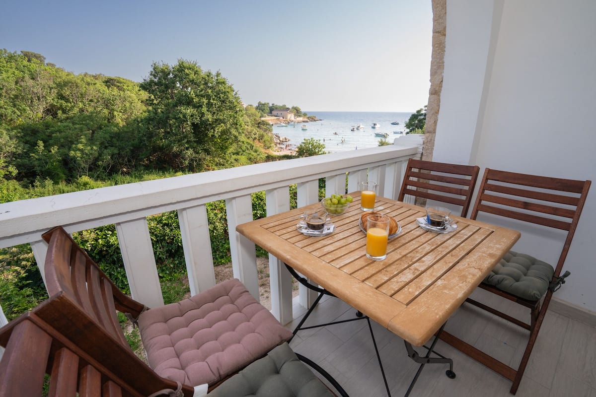 Un balcon avec une table dressée pour deux personnes, donnant sur une plage où l’on aperçoit des bateaux et une végétation luxuriante.