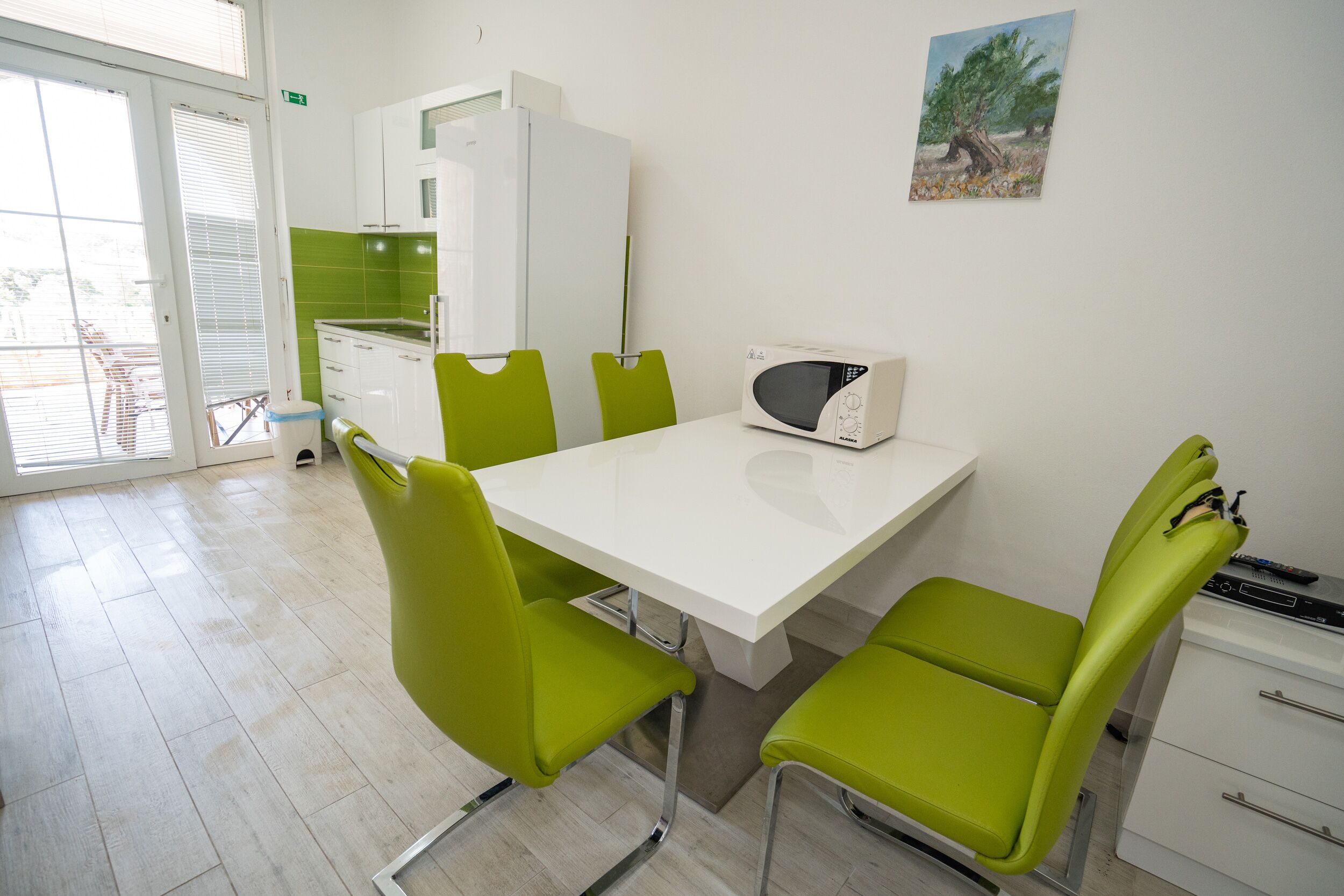 Un comedor con una mesa blanca, sillas verdes, un microondas y un cuadro en la pared.