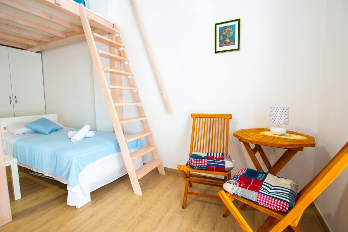 Une chambre avec un lit superposé, une échelle en bois, une chaise recouverte d’une courtepointe, une petite table avec une lampe et un tableau encadré au mur.