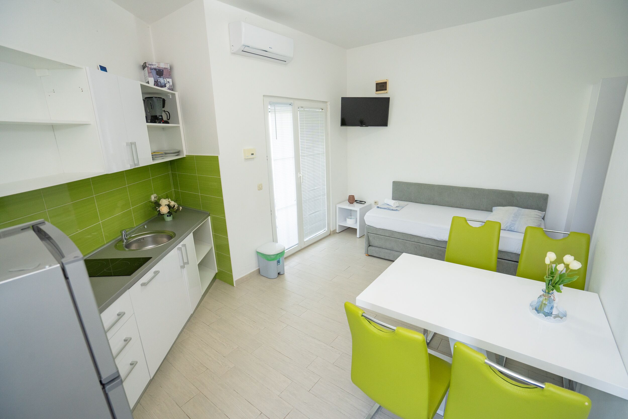 Habitación de hotel compacta con kitchenette, comedor con mesa y sillas, cama con cabecera gris y televisor montado en la pared.