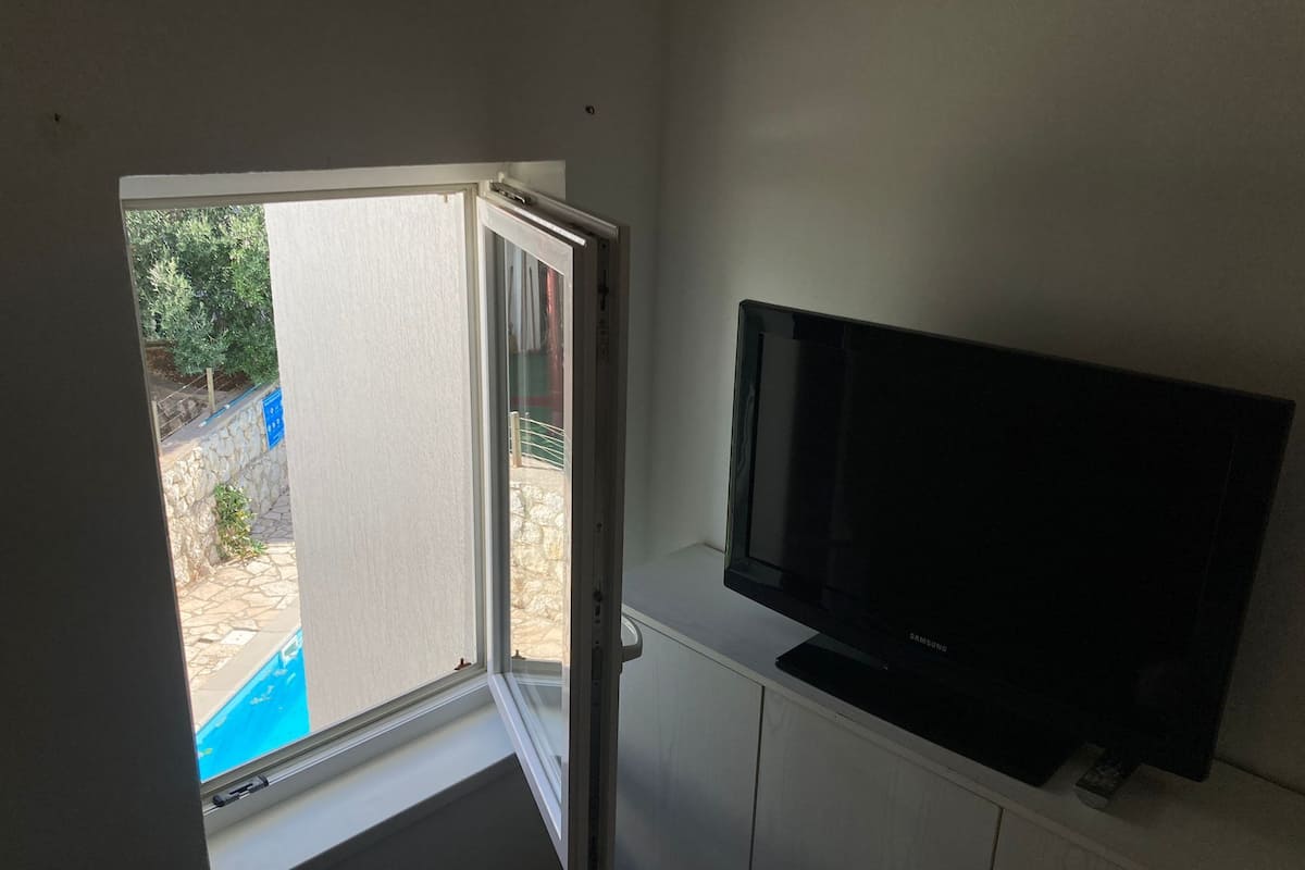 Une pièce avec un téléviseur posé sur un meuble, une fenêtre donnant sur une cour et une porte menant à l’extérieur.