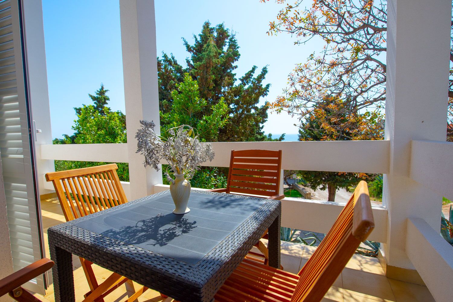 Een balkon met een tafel en stoelen, een vaas met bloemen, en uitzicht op bomen en de zee.