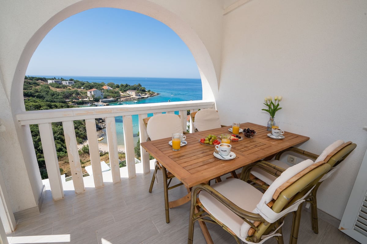 Une table en bois, entourée de chaises, sur un balcon offrant une vue sur la côte.