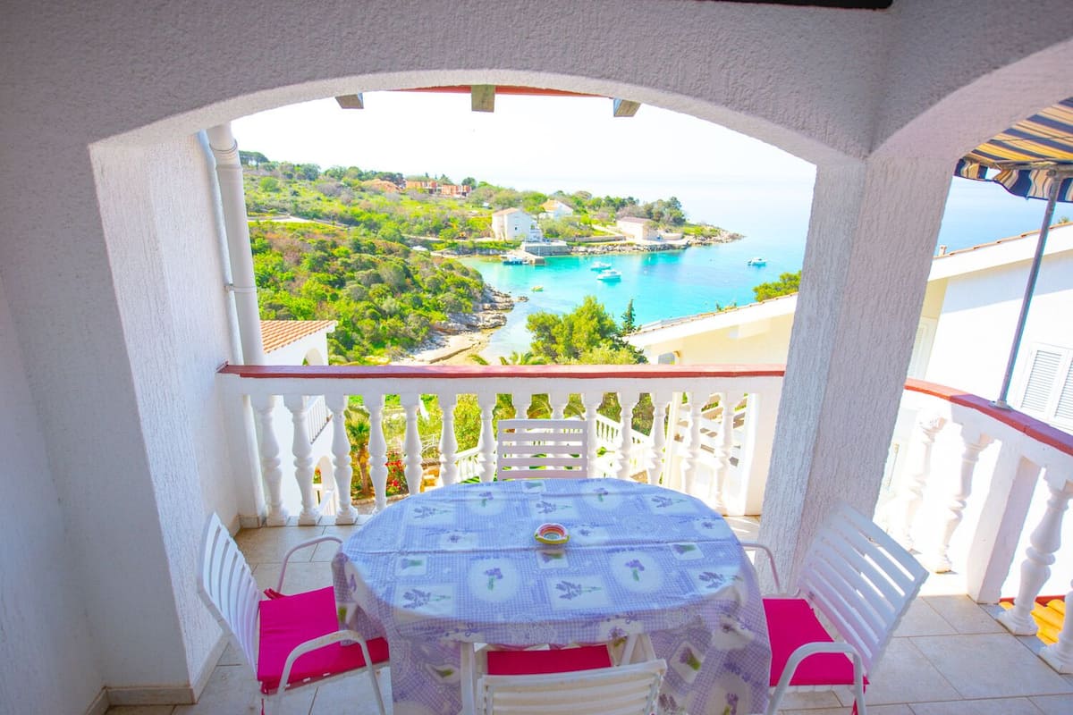 Un balcon avec une table et des chaises, donnant sur un paysage côtier avec des bâtiments et une plage.