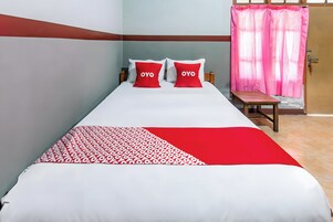 Una cama con ropa de cama blanca y roja, dos almohadas con el texto 'OYO', un banco de madera y una cortina rosa.