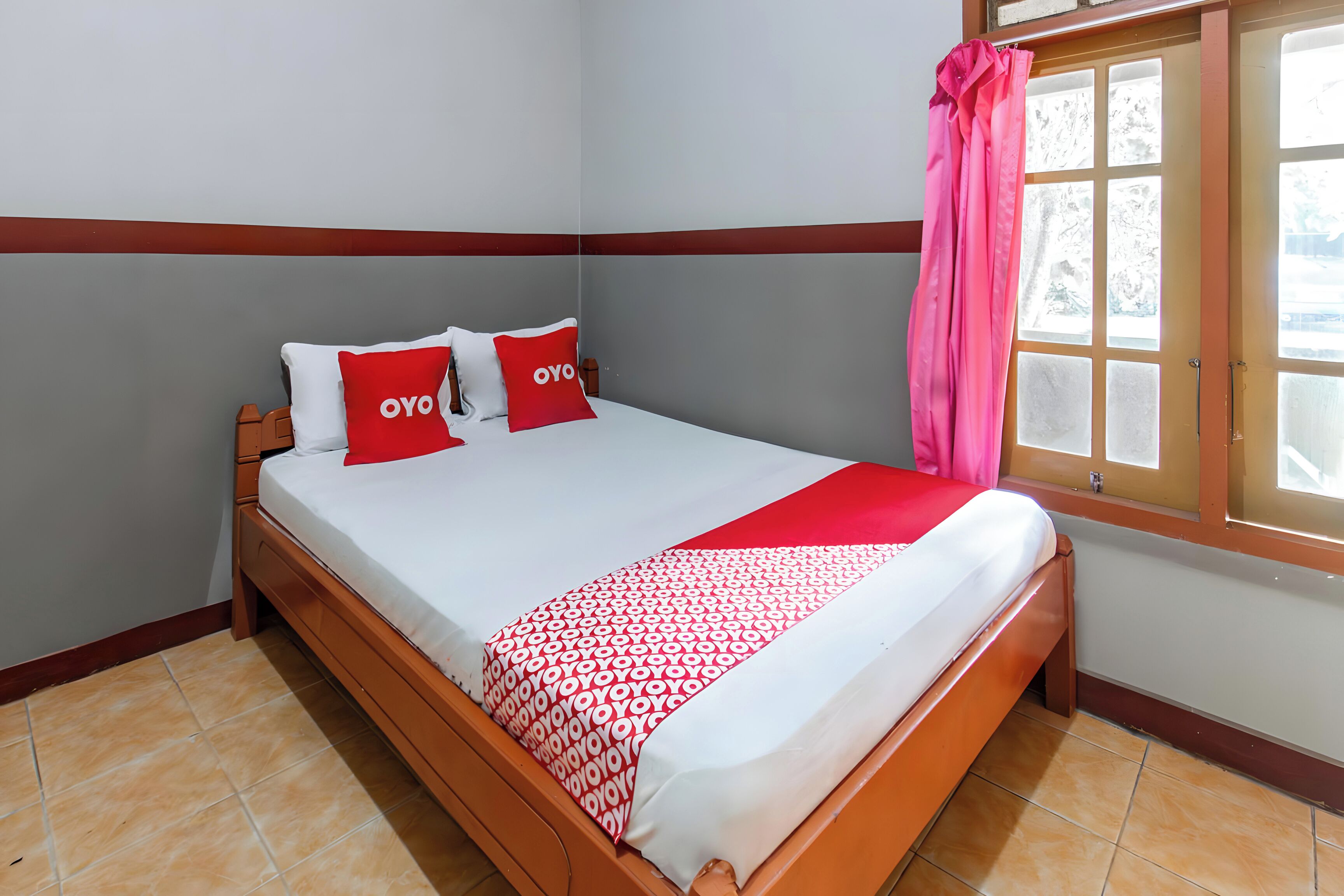 Una cama con una colcha blanca y roja estampada, dos almohadas con la palabra 'OYO' y una cortina rosa en una ventana.