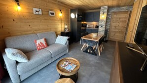 A cozy cabin interior with a wooden ceiling, a gray sofa with decorative pillows, a dining table with chairs, a small wooden coffee table, and a kitchen area with a microwave and stove.