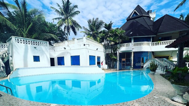 Una piscina con borde de baldosas, rodeada por un edificio blanco con ventanas azules y un balcón.