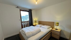 Une chambre à coucher avec un lit en bois, deux tables de chevet avec des lampes, une fenêtre avec des rideaux et une vue sur les arbres à l’extérieur.