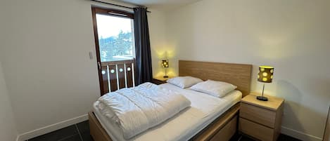Quarto com cama de madeira, duas mesas de cabeceira com abajures, uma janela com cortinas e vista para árvores lá fora.