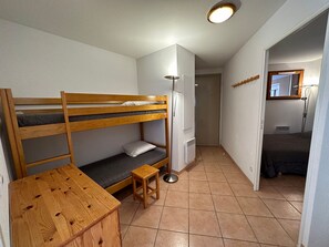 Ein kleines Zimmer mit Etagenbetten, einem kleinen Holztisch, einem Hocker, einer Heizung und einem Spiegel an der Wand.