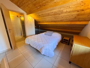 A bedroom with a bed, a wooden ceiling, a door leading to a bathroom, and a small wooden cabinet.