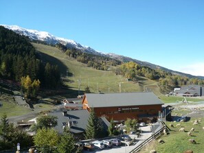 Uma pousada de montanha com um teleférico, rodeada por montanhas cobertas de neve e um estacionamento com carros.