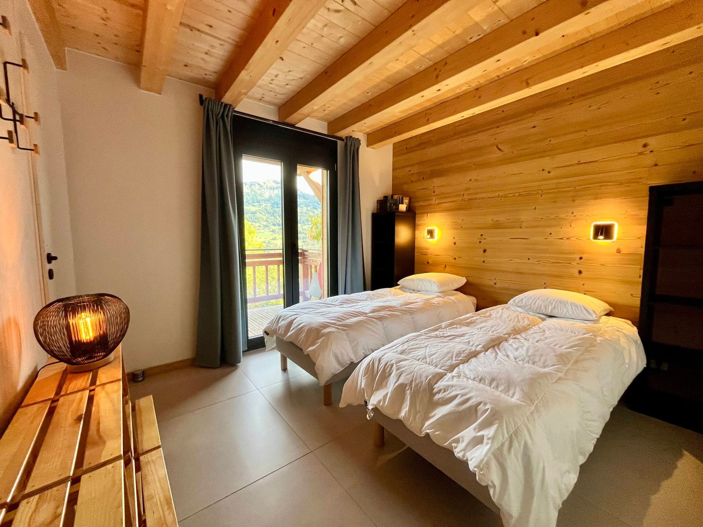 A bedroom with two beds, wooden ceiling beams, a balcony with a view, and a wall-mounted light fixture.