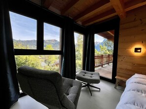 A room with a large window, a chair, a small stool, and a bed.
