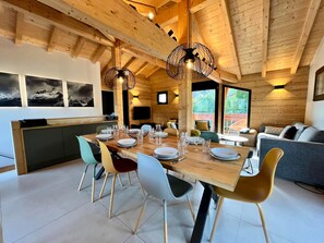 A modern interior with a wooden ceiling, a dining area with chairs and a table set, a sofa, and a television mounted on the wall.