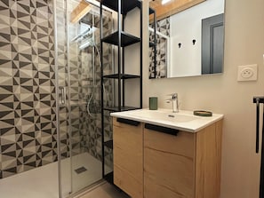 A modern bathroom with a geometric patterned shower wall, a glass-enclosed shower, a white sink with a single faucet, and a wooden vanity with cabinets.
