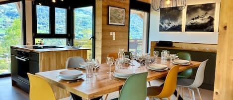 A dining area with a wooden table set for six, surrounded by colorful chairs, a kitchen with a view, and a dining area with artwork on the walls.