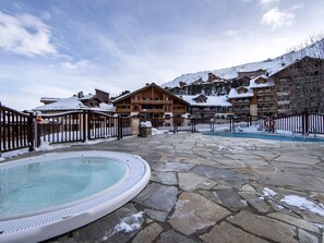 Ein Poolbereich mit Whirlpool, Steinboden, Holzhäusern und einem verschneiten Berg im Hintergrund.
