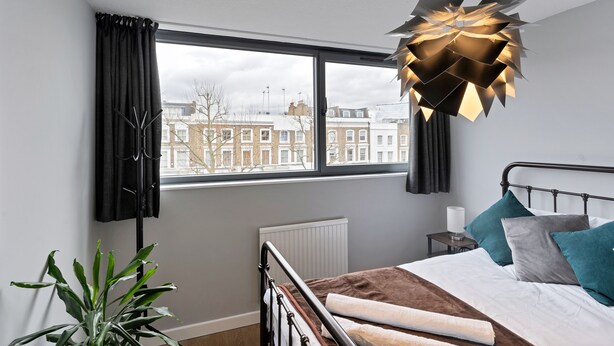 Une chambre moderne avec une grande fenêtre, un lit agrémenté de coussins turquoise et gris, un luminaire au design géométrique original et une plante en pot.