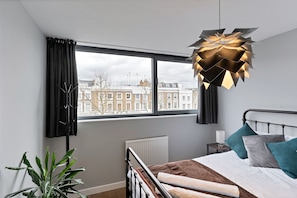 A modern bedroom with a large window, a bed with teal and gray pillows, a unique geometric light fixture, and a potted plant.