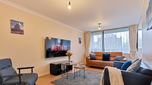 Un salon avec un téléviseur affichant le logo « NETFLIX », un canapé, un fauteuil, une table basse et une grande fenêtre avec des rideaux.