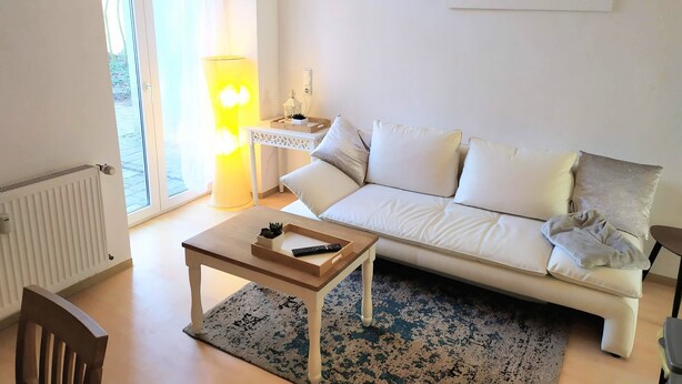 Ein Wohnzimmer mit einem weißen Sofa, einem Holztisch, einer Lampe und Blick durch eine Glastür.
